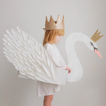 Una niña pequeña de perfil, con un vestido blanco y una corona dorada brillante, lleva un disfraz de cisne blanco con alas de papel y un pico rosado. El cisne también tiene una corona dorada. El fondo es blanco y minimalista, resaltando la escena.